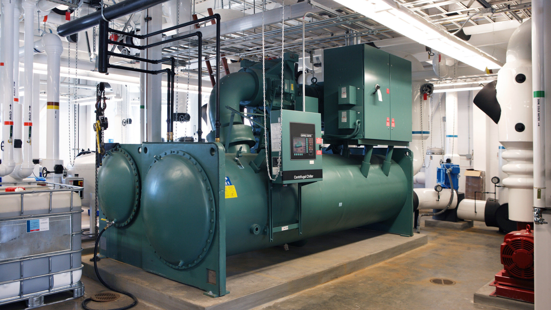Large industrial chiller unit with control panel, surrounded by pipes and ducts in a mechanical room. Large industrial chiller unit with control panel, surrounded by pipes and ducts in a mechanical room.