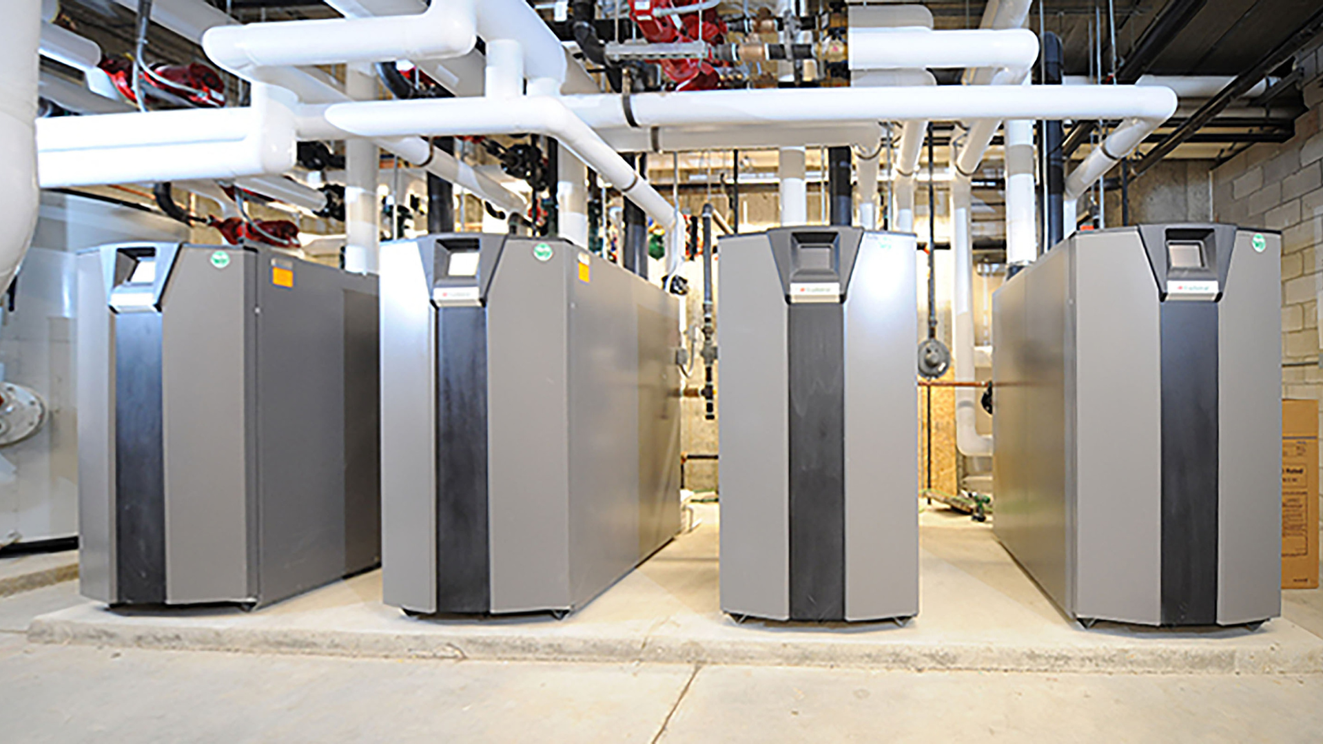 Four large commercial HVAC units are installed on a concrete floor, surrounded by white and red pipes in an industrial utility room. Four large commercial HVAC units are installed on a concrete floor, surrounded by white and red pipes in an industrial utility room.