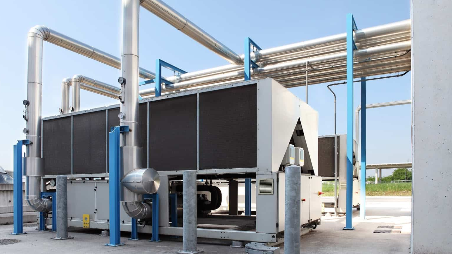 Large industrial HVAC chiller unit with connected metal pipes and support structures, located outdoors on a concrete platform under a clear sky. Large industrial HVAC chiller unit with connected metal pipes and support structures, located outdoors on a concrete platform under a clear sky.