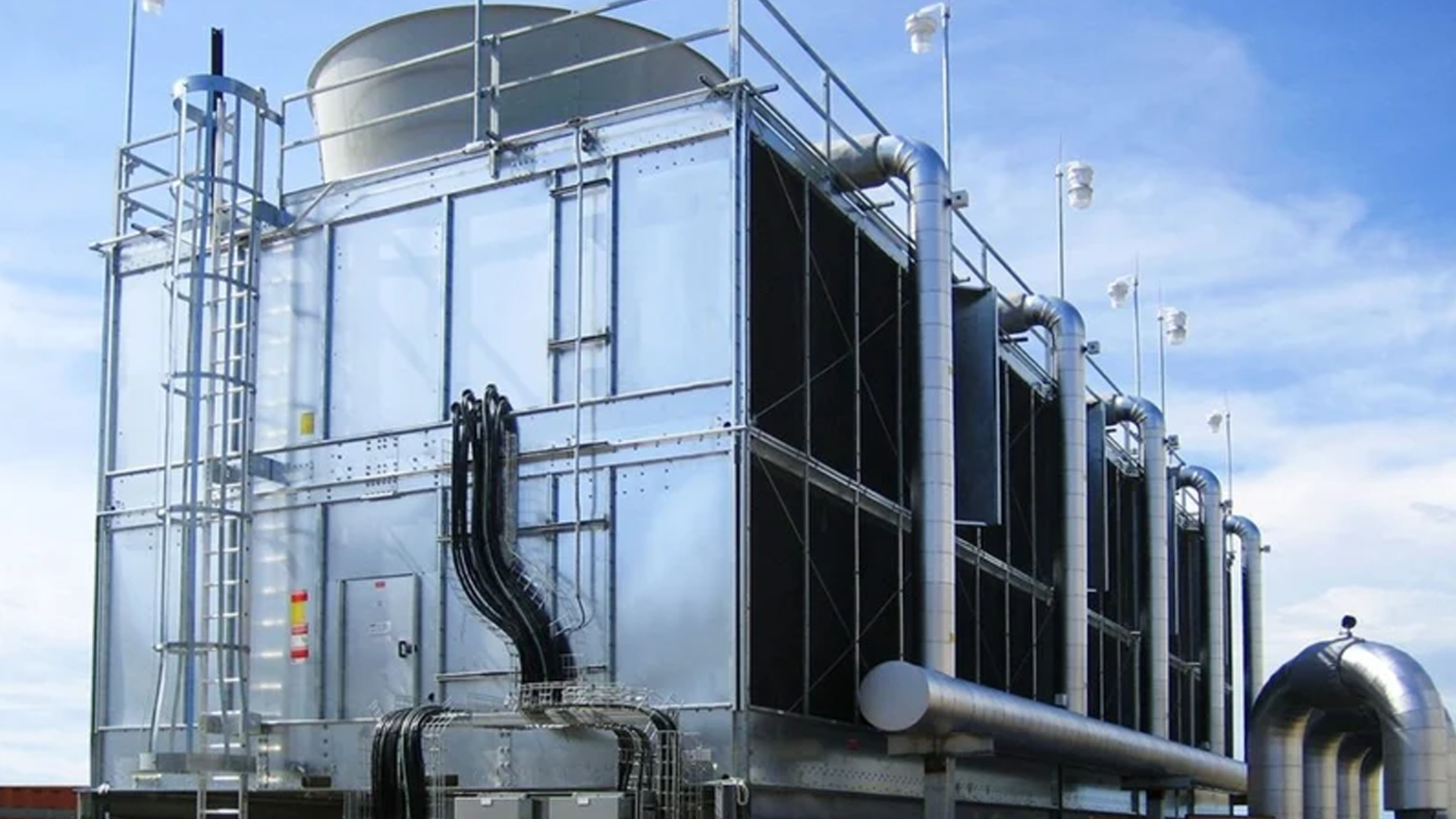 Large industrial cooling tower with metal exterior, multiple pipes, vents, and electrical conduits, shown outdoors under a partly cloudy sky. Large industrial cooling tower with metal exterior, multiple pipes, vents, and electrical conduits, shown outdoors under a partly cloudy sky.