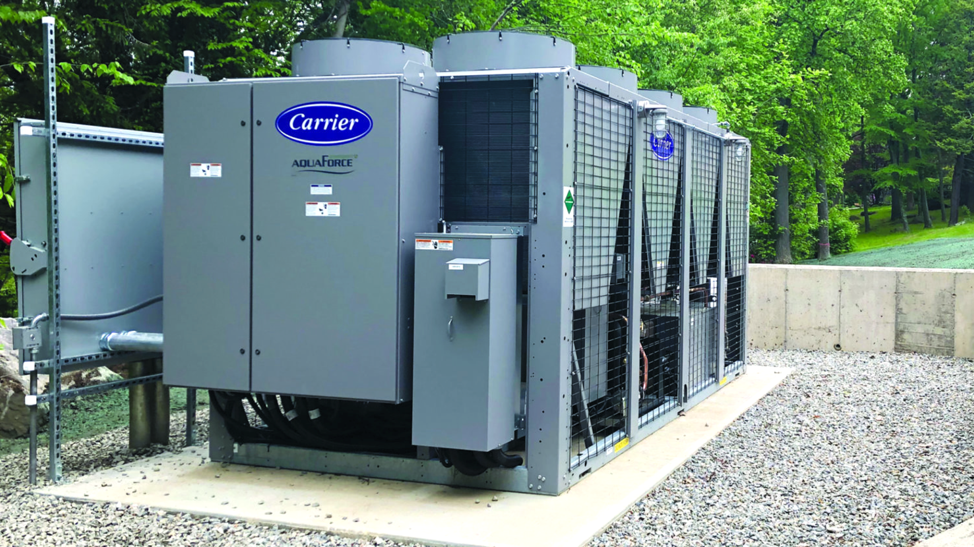A Carrier AquaForce industrial HVAC unit is installed outdoors on a gravel pad, surrounded by trees and a concrete retaining wall. A Carrier AquaForce industrial HVAC unit is installed outdoors on a gravel pad, surrounded by trees and a concrete retaining wall.