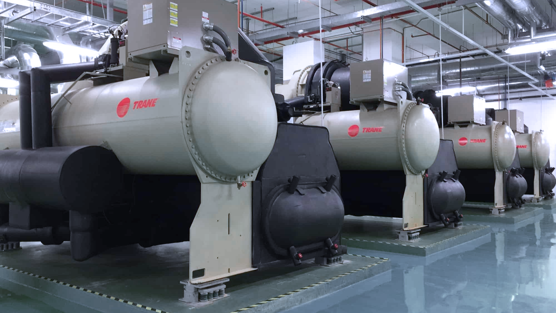 Several large industrial Trane chillers are installed in a clean, well-lit mechanical room with reflective flooring and overhead piping. Several large industrial Trane chillers are installed in a clean, well-lit mechanical room with reflective flooring and overhead piping.