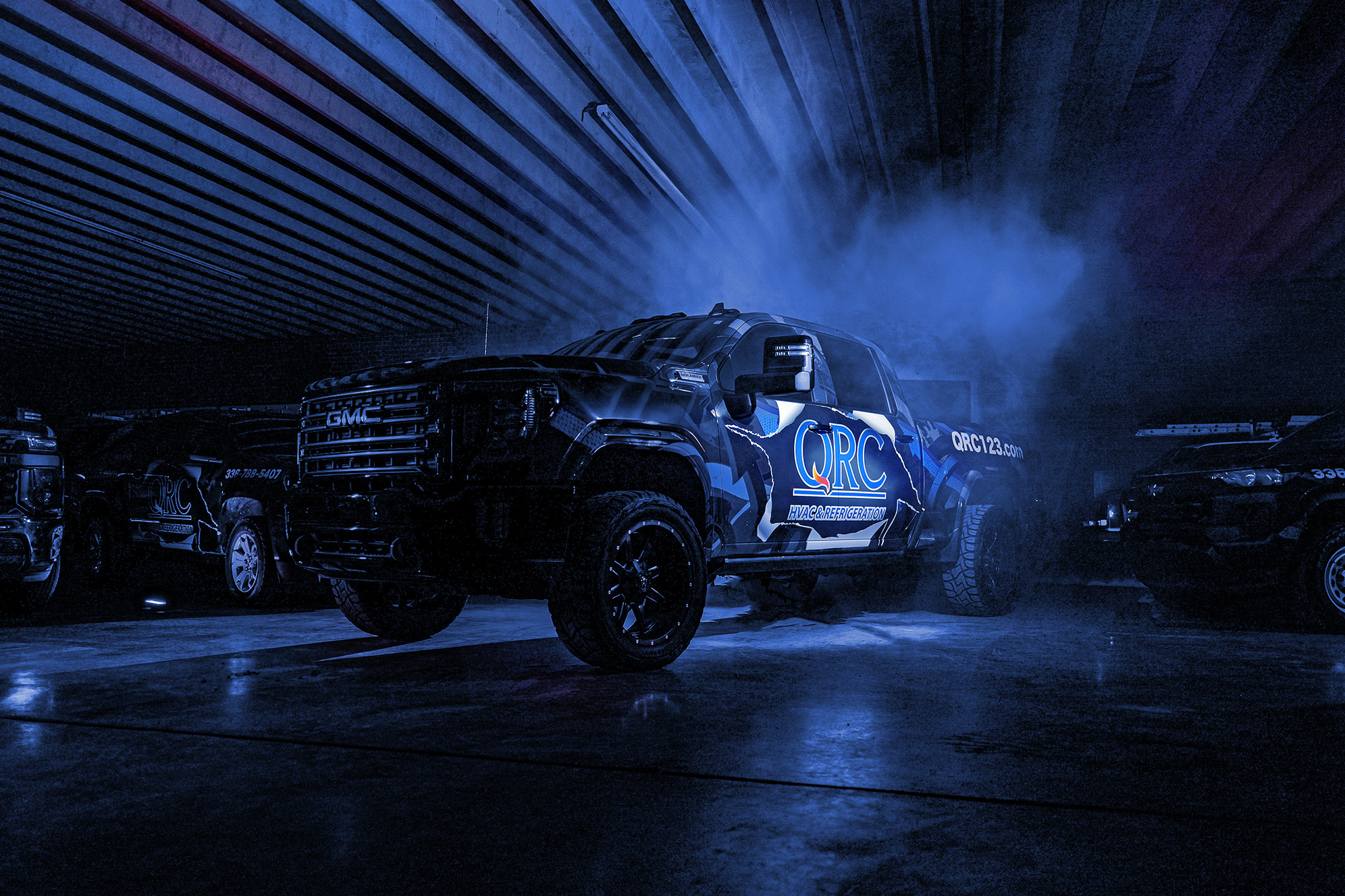 A GMC pickup truck with QRC branding is parked indoors under blue lighting, with smoke or mist visible around it. A GMC pickup truck with QRC branding is parked indoors under blue lighting, with smoke or mist visible around it.