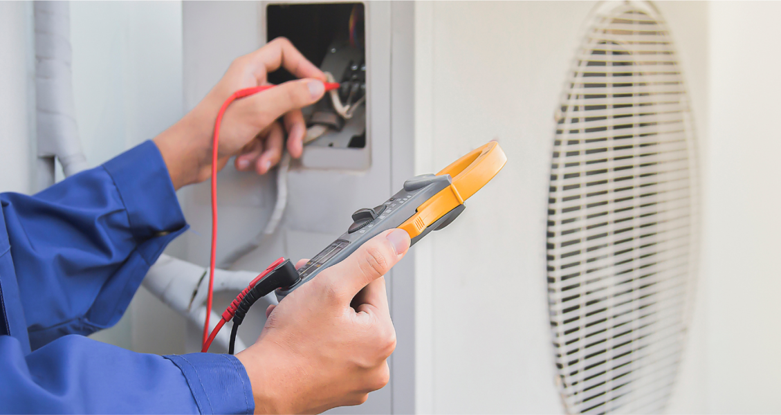A person in a blue sleeve uses a digital clamp meter to test wiring on an air conditioning unit. A person in a blue sleeve uses a digital clamp meter to test wiring on an air conditioning unit.