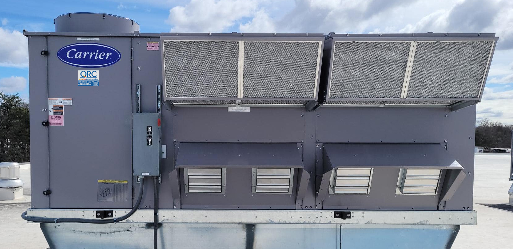 Large rooftop Carrier HVAC unit with open filter panels, electrical box on side, and vent openings, set against a partly cloudy sky.